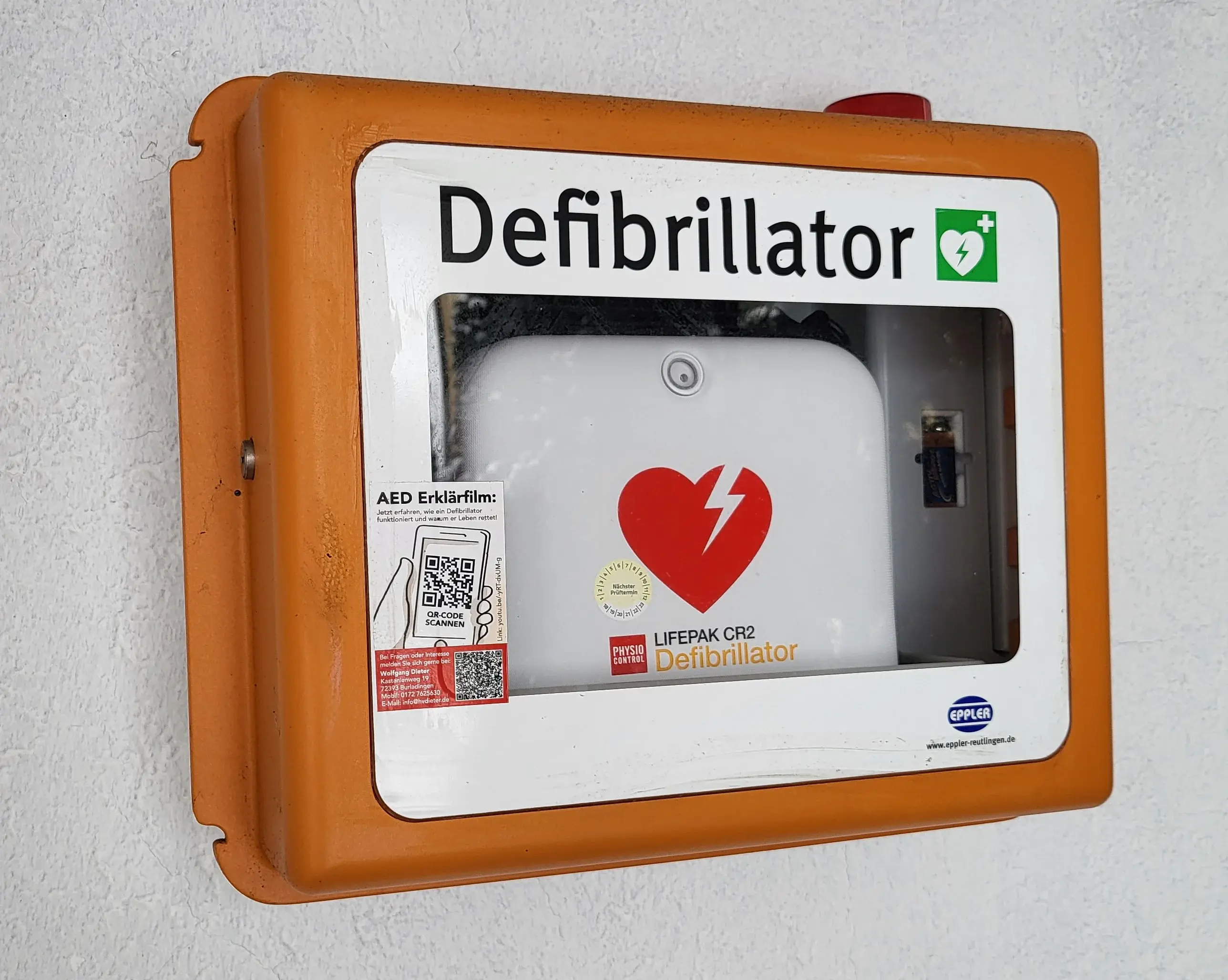 Auf dem Bild ist der Defibrillator in einem orange-weißen Kasten zu sehen. In der Mitte ist ein rotes Herz auf dem weißen Gerät. Es steht Defibrillator drauf und daneben ist ein kleines grünes Quadrat, darin ist ein weißes Herz mit einem grünen Pfeil und oben rechts in diesem kleinen grünen Quadrat ist noch ein weißes Kreuz darin. Dieses ist das Symbol für den Defibrillator