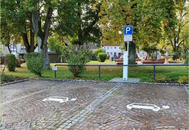 Auf dem Bild sieht man den Parkplatz mit jeweils zwei Flächen darauf, auf denen ist jeweils ein in weiß bemaltes Auto mit einem Ladekabel als Symbol für hier kann geladen werden, drauf. Im Hintergrund sind die grünen Bäume des Parkes zu sehen wie auch ein Schild in blau mit einem weißen Buchstaben P darauf.